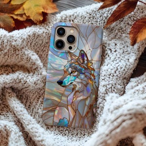 May include: A phone case featuring a stained-glass style wolf design. The case has a pearlescent finish with blue, gold, and white tones. The wolf is depicted in profile, with intricate details. The case is resting on a soft, textured surface.