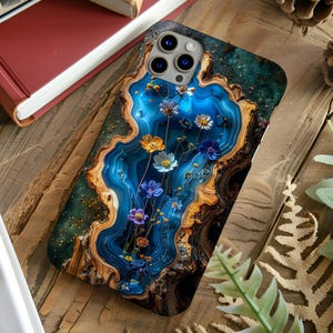 May include: A rectangular phone case featuring a vibrant design of blue agate stone patterns with concentric rings of tan and brown wood grain. The center displays a vertical arrangement of delicate wildflowers in shades of blue, orange, and white.