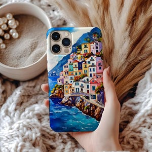 May include: A smartphone case featuring a colorful watercolor painting of a coastal Italian village. The design includes buildings in shades of pink, yellow, and blue, set against a backdrop of a blue sea and sky. The case has a glossy finish.