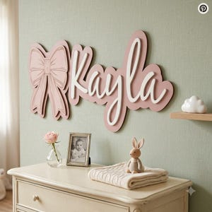 May include: A wall-mounted sign featuring the name Kayla in white script lettering layered over a dusty pink backing with a large bow icon on the left side. The sign is displayed above a dresser with a small vase, a photo frame, and a toy.
