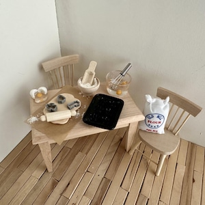 Könnte beinhalten: Ein Miniatur-Holztisch mit zwei Stühlen, ausgestattet mit Backzubehör wie Nudelholz, Ausstechformen, einer Schüssel mit Holzlöffel, einem Schneebesen in einer Glasschüssel mit Ei, einem schwarzen Backblech und einem weißen Beutel mit Aufschrift Old Mill Flour 50LBS.