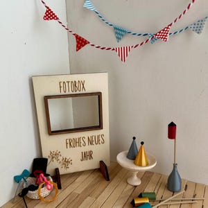 Könnte beinhalten: Eine Miniatur-Partyszene mit einer Holz-Fotobox mit der Aufschrift "FOTOBX" und "FROHES NEUES JAHR". Enthält Partyhüte, Requisiten und ein Banner mit roten, weißen und blauen Wimpeln. Ein kleiner Tisch hält zwei Partyhüte.