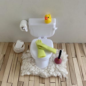 Könnte beinhalten: Ein Miniatur-Toilettenset in Weiß mit einer gelben Quietscheente auf dem Spülkasten, hellgrünen Putzhandschuhen auf dem Sitz, einem kleinen Pümpel, einer Rolle Toilettenpapier in einem geflochtenen Korb und einem Wand-Toilettenpapierhalter.