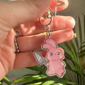 May include: Pink bunny keychain with a silver star charm and a small knife. The bunny is holding the knife in its right hand.