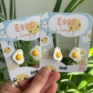 Peut inclure: Deux paires de boucles d'oreilles en forme d'œuf blanc et jaune avec des crochets argentés. Les boucles d'oreilles sont présentées sur une carte avec le texte "Eggs Earrings" et "Handmade with Love".