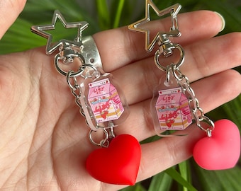 May include: Two pink and white milk carton charms with a red and pink heart charm attached to each. The charms are attached to silver star-shaped keychains.