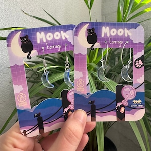 Puede incluir: Un par de pendientes de media luna con un gato negro en una media luna en el embalaje. El embalaje es morado y azul con el texto "Mook Earrings" y "Handmade with Love".
