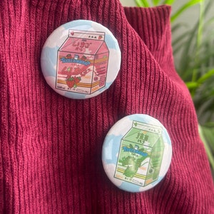 Puede incluir: Dos pines redondos con ilustraciones de dibujos animados de cajas de leche. El pin rosa presenta una caja de leche de fresa con el texto "いちごみるく" y el pin verde presenta una caja de leche de té verde con el texto "抹茶みるく".