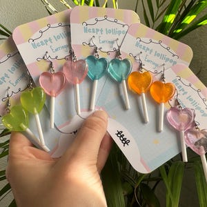 Puede incluir: Una mano sostiene varios pares de pendientes de piruleta en forma de corazón en varios colores: verde, rosa, azul y naranja. Cada pendiente está unido a un palito blanco y se muestra en una tarjeta de color pastel con el texto "Heart lollipop Earrings".