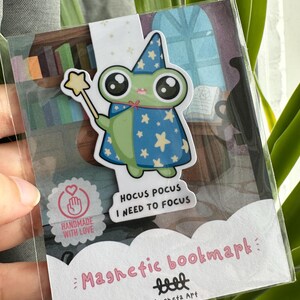 Puede incluir: Marcapáginas magnético con un diseño de rana mago. La rana lleva una capa azul con estrellas y sostiene una varita mágica. El texto en el marcapáginas dice "Hocus Pocus I Need to Focus" y "Magnetic bookmark" en rosa. También se ve "Handmade with Love".