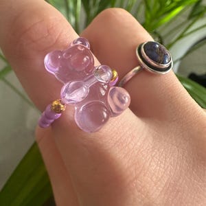 Puede incluir: Primer plano de dos anillos. Uno presenta un diseño de oso de peluche morado translúcido con detalles dorados. El otro anillo tiene una banda plateada y una piedra ovalada azul oscuro. El fondo está borroso con follaje verde.