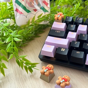 Puede incluir: Teclas de teclado con forma de hombre de jengibre con pajaritas rojas y detalles azules se muestran en un teclado con teclas lavanda y negras. Una tecla también está empaquetada en una funda de plástico transparente con un respaldo a rayas rojas, blancas y verdes.