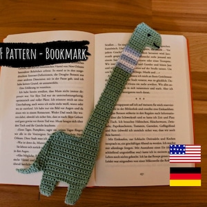 Könnte beinhalten: Ein grünes, gehäkeltes Dinosaurier-Lesezeichen mit einem weiß-blau gestreiften Hals. Das Lesezeichen liegt auf einem geöffneten Buch mit dem sichtbaren Text "PDF PATTERN - BOOKMARK". Das Bild zeigt auch die amerikanische und deutsche Flagge.