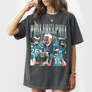 Puede incluir: Una camiseta gris carbón de manga corta con un estampado gráfico de estilo vintage. El diseño muestra el texto PHILADELPHIA FOOTBALL sobre un collage de cuatro jugadores de fútbol americano con uniformes verde azulado, incluyendo los números 26, 11.