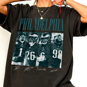 Puede incluir: Una camiseta gris carbón con la palabra PHILADELPHIA en texto verde azulado sobre cuatro paneles que muestran jugadores de fútbol americano con uniformes oscuros. Cada panel muestra a un jugador con casco y camiseta con los números 1, 26, 6 y 98, con firmas.