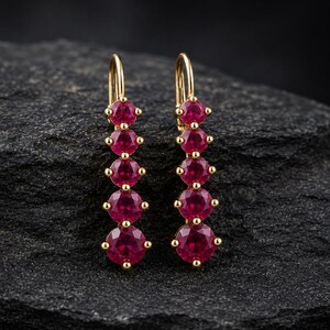 Puede incluir: Un par de pendientes colgantes con cinco rubíes redondos de color rosa intenso engastados en oro amarillo. Cada pendiente presenta una hilera vertical de piedras sujetas por pequeñas garras, suspendidas de un gancho de oro curvado.
