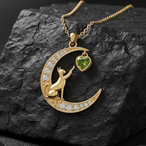 May include: A gold necklace with a crescent moon pendant set with clear stones. A cat silhouette reaches for a heart-shaped green stone. The necklace is displayed on a dark, textured surface.