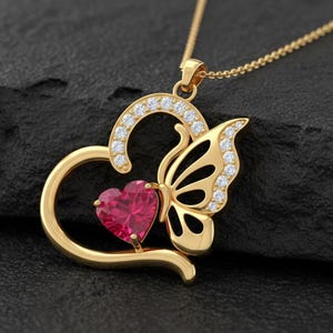 Puede incluir: Un collar con colgante dorado con un colgante en forma de corazón con una piedra preciosa roja y un diseño de mariposa. El colgante está adornado con pequeñas piedras preciosas transparentes. El collar se presenta sobre una superficie oscura.