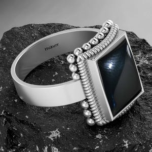 May include: A silver ring with a large, square, black gemstone. The ring has a wide band and a decorative border of small silver beads around the gemstone. The word "Huzurr" is engraved on the inside of the band. The ring is displayed on a dark, textured surface.