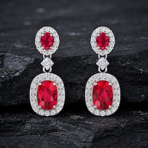 Puede incluir: Un par de pendientes colgantes con gemas rojas ovaladas rodeadas de pequeñas piedras blancas. Cada pendiente tiene una piedra roja ovalada más pequeña en la parte superior y una más grande en la parte inferior, engastadas en metal plateado.