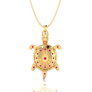 Könnte beinhalten: Eine goldfarbene Schildkröten-Anhänger-Halskette mit einer filigranen Kette. Der Schildkröten-Anhänger hat ein detailliertes Design mit roten Edelstein-Akzenten. Die Halskette ist ein Schmuckstück, geeignet zum Verschenken oder Tragen.