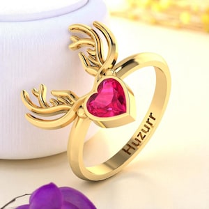 Pode incluir: Anel dourado com uma pedra preciosa rosa brilhante em forma de coração. O anel apresenta um design de chifres de veado. A palavra "Huzurr" está gravada na banda do anel. Uma joia decorativa.