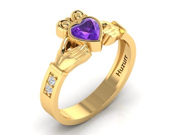 May include: A gold ring with a purple heart-shaped gemstone and two small diamonds on the band. The ring is a Claddagh ring, a traditional Irish design featuring two hands clasping a heart.