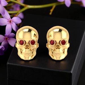 May include: A pair of gold-colored skull-shaped earrings with red gemstone eyes. The earrings are displayed on a black surface, with purple flowers in the background. The earrings have a polished finish.