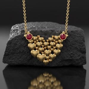 Puede incluir: Un collar dorado con un colgante en forma de corazón compuesto por numerosos corazones pequeños. El collar tiene una cadena fina y está adornado con dos gemas rojas redondas. La joya se exhibe sobre una superficie oscura y texturizada.