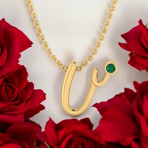 May include: A gold necklace with a cursive letter 'V' pendant and a small green gemstone. The necklace is on a white background with red roses.