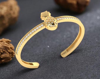 May include: A gold-colored open cuff bracelet with a pineapple-shaped design. The bracelet is encrusted with small, clear gemstones along the top and around the pineapple. The word "Huzurr" is engraved on the inside of the bracelet.
