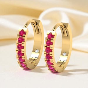 Könnte beinhalten: Ein Paar goldfarbene Creolen. Jeder Ohrring hat eine Reihe leuchtend rosa Edelsteine und einen kleinen, klaren Akzentstein. Die Ohrringe haben eine polierte Oberfläche und ein klassisches Design.