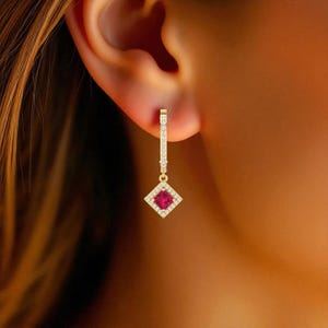 May include: A gold earring with a rectangular red gemstone center, surrounded by small white gemstones. The earring is suspended from a gold bar with small white gemstones. The earring is shown on a person's ear.