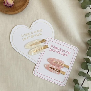 May include: Two sets of hair clips on decorative cards. One set features white and cream-coloured clips, the other pink and white. The cards have the text "To have & to hold your hair back" in a script font.