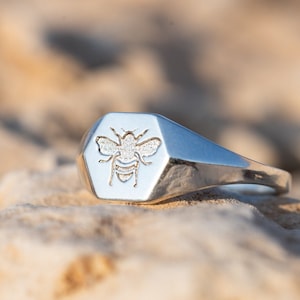 May include: A silver signet ring with a hexagonal face featuring an engraved bee design. The ring is resting on a light brown textured surface.