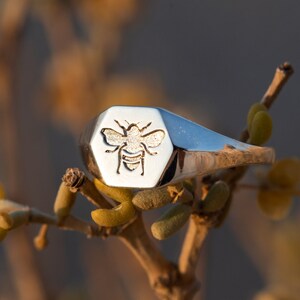 May include: A silver signet ring with a hexagonal face featuring an engraved bee design.