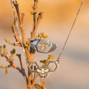 May include: A silver necklace with a bee and honeycomb pendant hangs from a branch. A silver ring with a bee design is also visible.