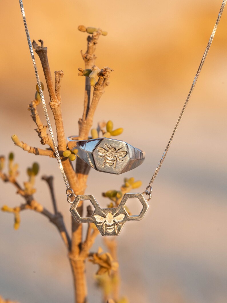 May include: A silver necklace with a bee and honeycomb pendant hangs from a branch. A silver ring with a bee design is also visible.