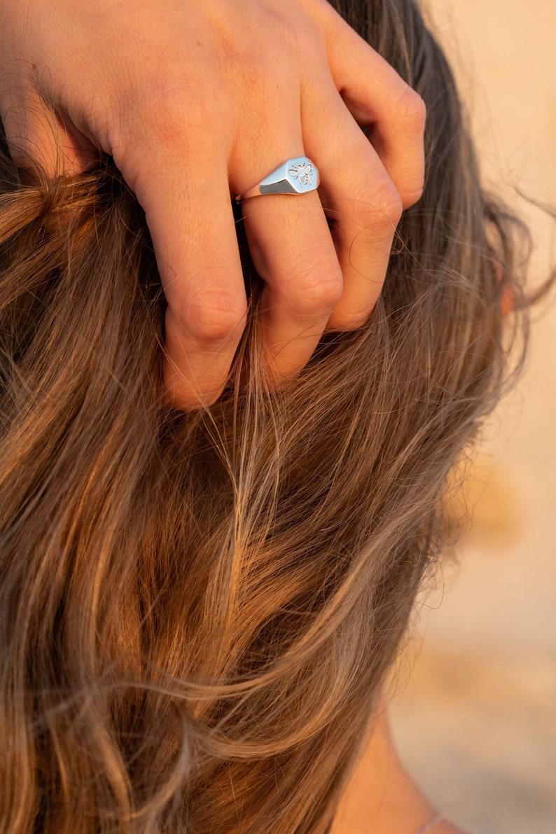 May include: A silver signet ring with a bee design. The ring is worn on the finger of a person with long brown hair.
