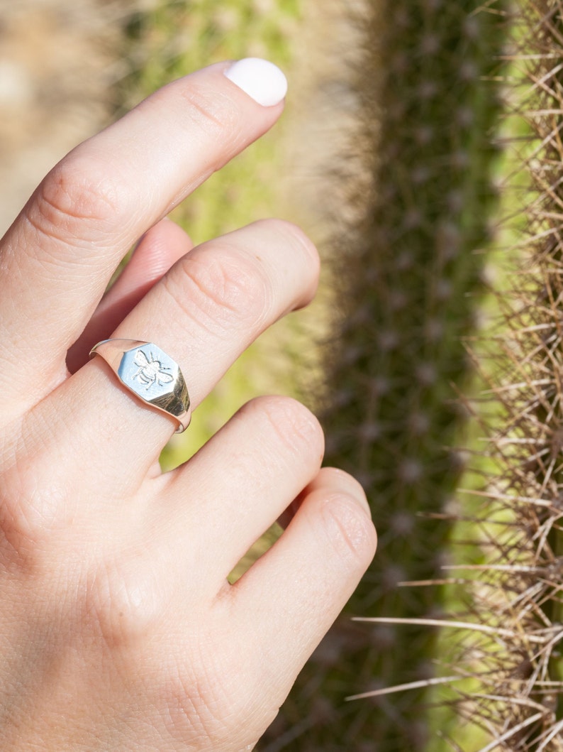 May include: A silver signet ring with a bee design. The ring is worn on a person's finger.