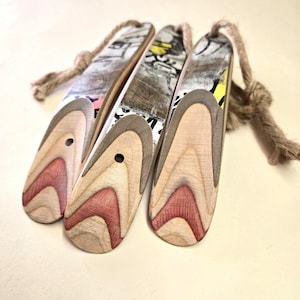 Könnte beinhalten: Drei Schuhanzieher aus Holz mit natürlicher Holzmaserung und einem geschichteten Design in Rot, Braun und Beige. Die Oberseiten haben ein abgenutztes Graffiti-Design und sind an einer Naturkordel befestigt. Jedes Horn hat ein kleines Loch.