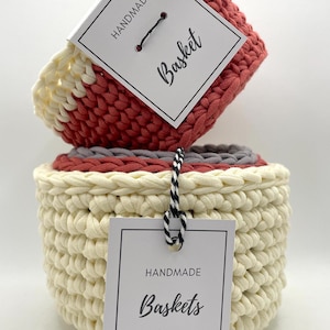 Könnte beinhalten: Drei gehäkelte Körbe in verschiedenen Größen, übereinander gestapelt. Der obere Korb ist rot und weiß, der mittlere Korb ist grau und weiß und der untere Korb ist cremefarben. Die Körbe sind handgefertigt und haben ein Etikett mit der Aufschrift "Handgefertigte Körbe".
