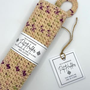 May include: A handmade beige pot holder with pink and green accents. The pot holder has a loop for hanging and a tag that reads "I'm a handmade pot holder that makes me one of a kind 100% cotton".