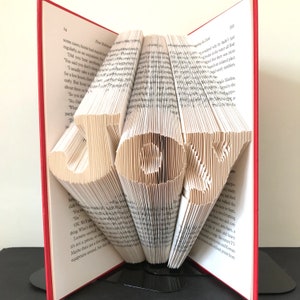 May include: A book with the word "JOY" folded into the pages. The book is open and the pages are folded to create the word. The book is on a black stand.