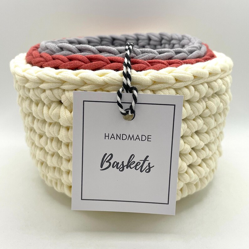 Pode incluir: Conjunto de tr&ecirc;s cestas de croch&ecirc; em creme, vermelho e cinza. As cestas est&atilde;o empilhadas umas sobre as outras. Uma etiqueta branca com texto preto diz "HANDMADE Baskets".