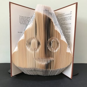 May include: A book folded into the shape of a face with two large eyes. The book is open and the pages are folded to create the shape of the face. The book is standing on a black surface.