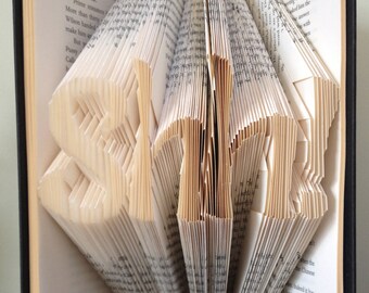 May include: A book art sculpture of the word "SHH" created by folding the pages of a book. The book is open and the pages are folded to create the letters. The book is sitting on a black surface.