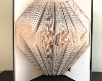 May include: A book with the word "HOME" folded into the pages. The book is open and the pages are folded to create a 3D effect. The book is on a black surface.