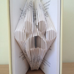 May include: A book folded into a three-dimensional heart shape. The book is open and the pages are folded to create the heart shape. The text on the pages is visible, but it is difficult to read. The book is sitting on a wooden surface.