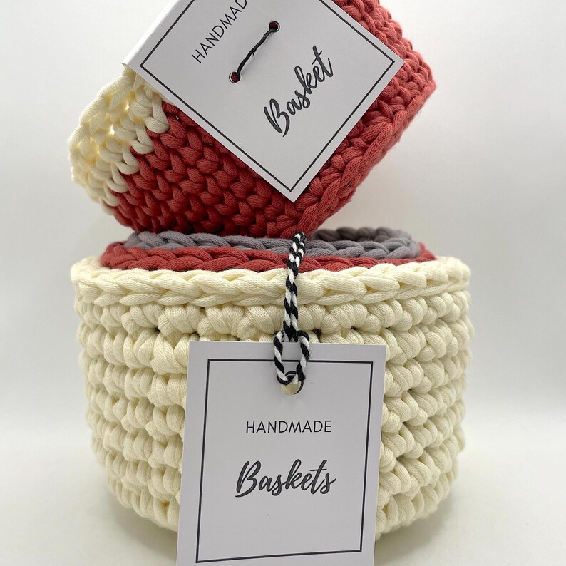 Pode incluir: Tr&ecirc;s cestas de croch&ecirc; de tamanhos diferentes, empilhadas umas sobre as outras. A cesta superior &eacute; vermelha e branca, a cesta do meio &eacute; cinza e branca, e a cesta inferior &eacute; creme. As cestas s&atilde;o feitas &agrave; m&atilde;o e t&ecirc;m uma etiqueta que diz "Cestas feitas &agrave; m&atilde;o".